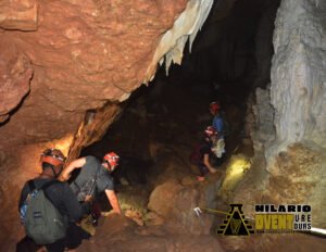 Two smiling women wearing helmets and harnesses stand in a lit limestone cave surrounded by stalactites—their headlamps glowing softly in the underground space with Hilario Adventure Tours